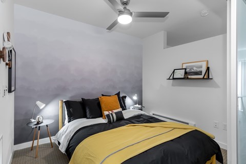 A bedroom with a bed, a ceiling fan, and a small table with a lamp.