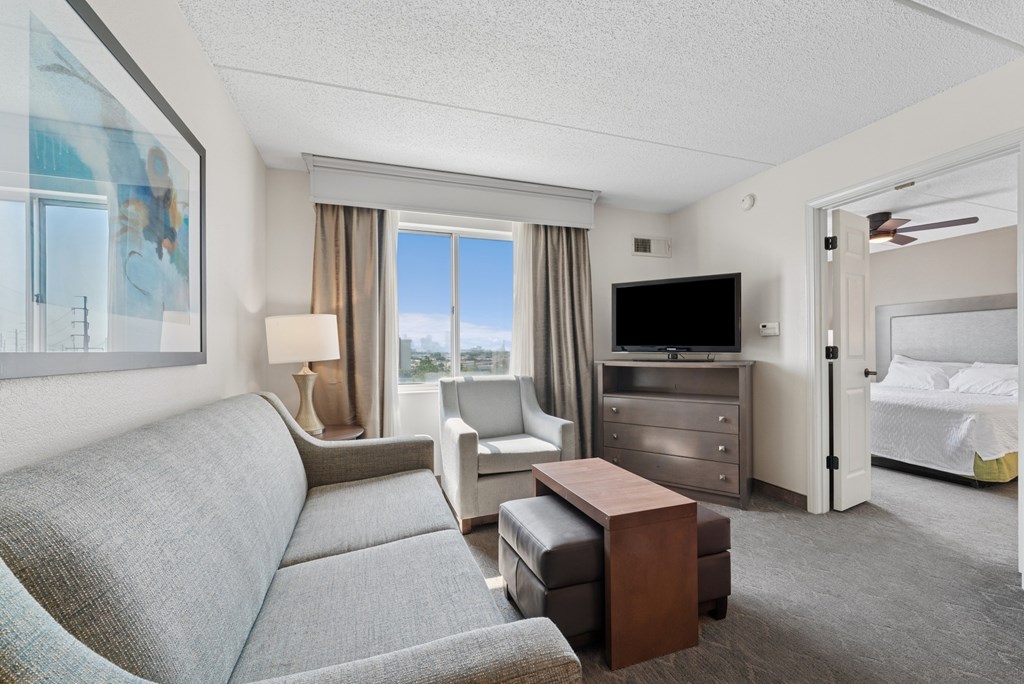 A well-lit hotel room with a grey sofa, a bed, a desk, and a television.