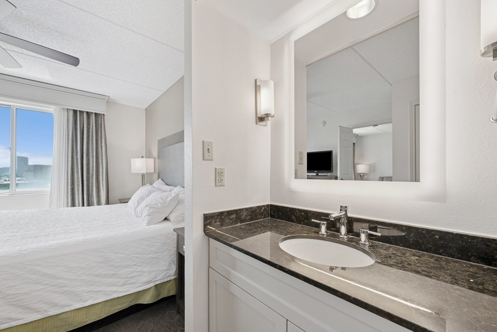 A well-lit hotel bathroom with a large mirror and a bed with white bedding.