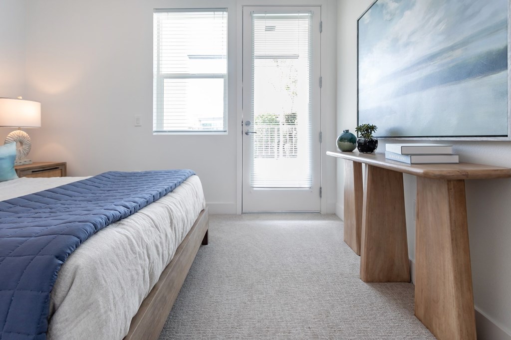 A bedroom with a bed, a nightstand, a lamp, a table with a plant and a painting on the wall.