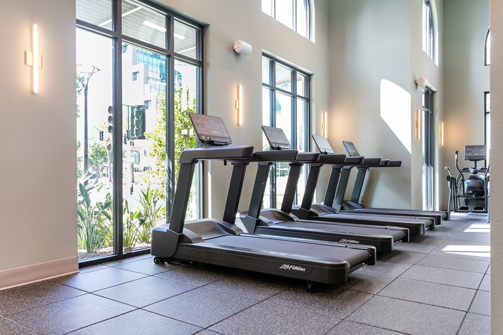 A row of treadmills are lined up in a room with a view of the city outside.
