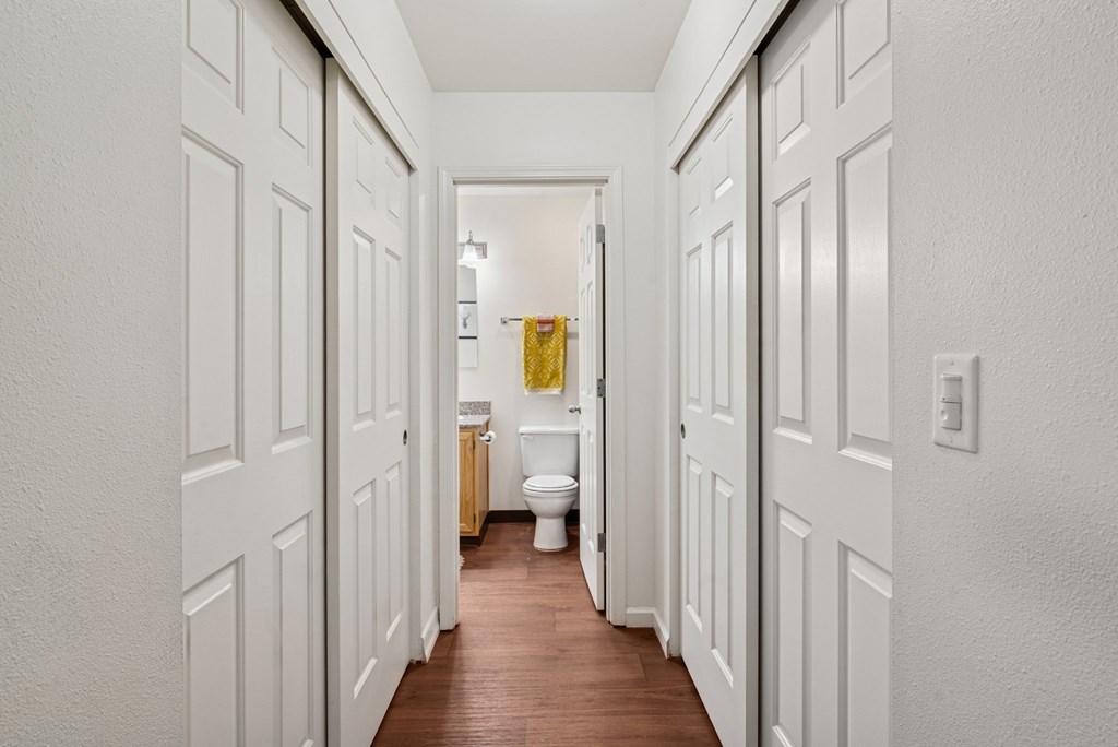A narrow hallway with white walls and a wooden floor leads to a bathroom.