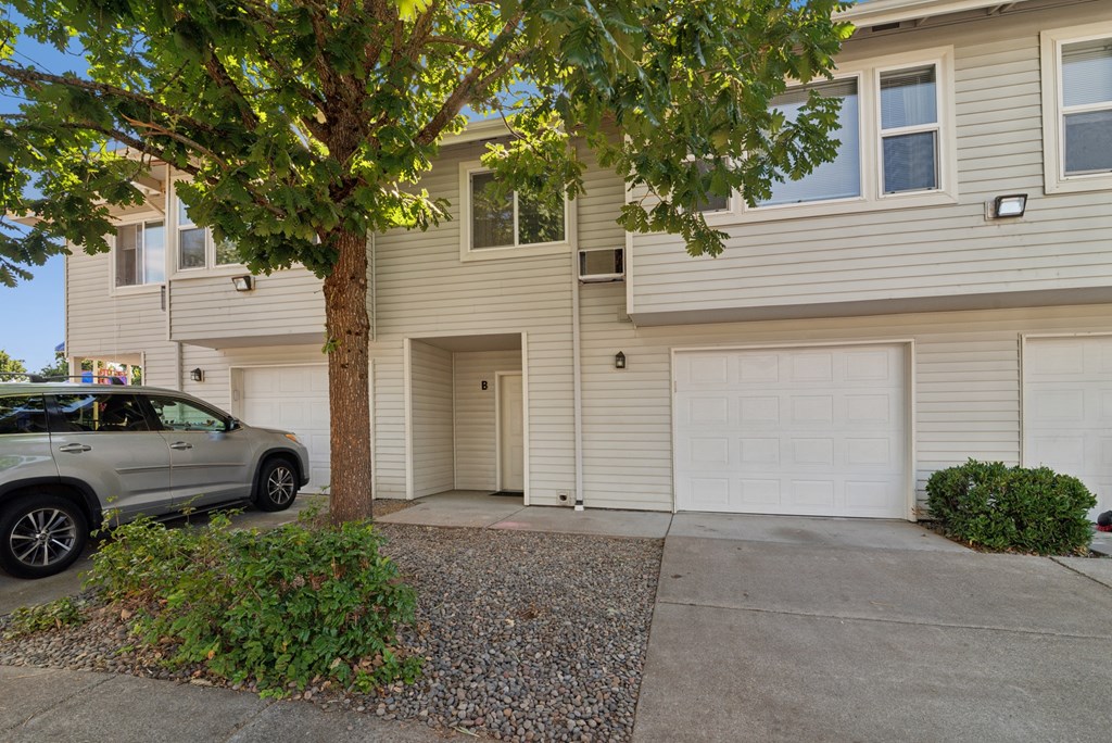 A grey SUV is parked in front of a two-story beige house with a tree in front.