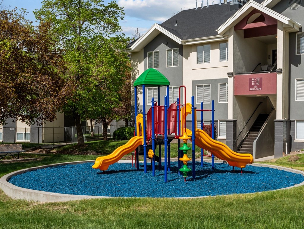 A playground with a green roof and a blue base is in front of a grey apartment building.