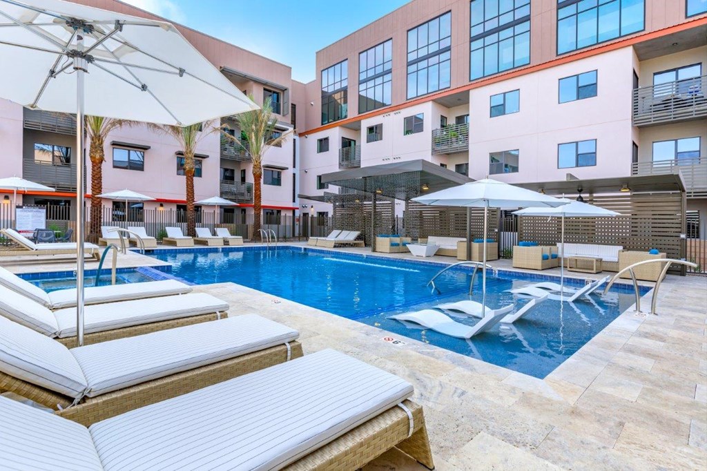 A pool area with sun loungers and umbrellas in front of a multi-story building.