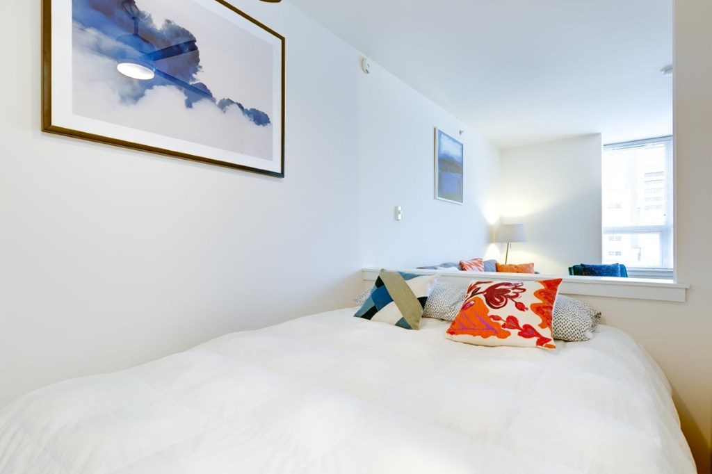 A bed with a white comforter and a blue pillow is in the foreground of a room with a framed picture of a blue sky with clouds on the wall.