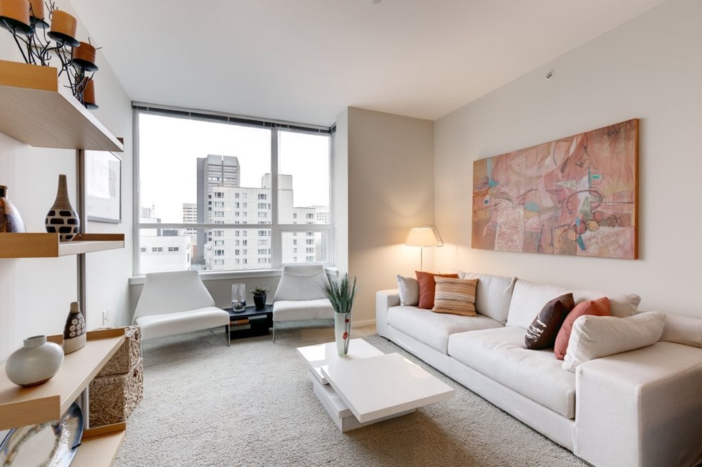 A living room with a white couch, a coffee table, and a large window overlooking a cityscape.