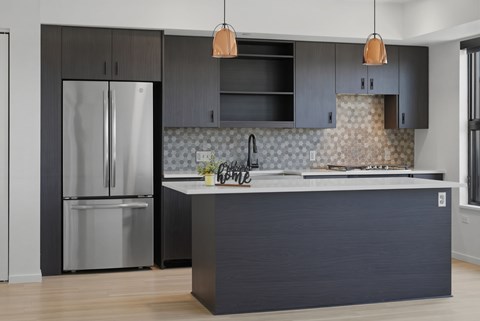 A modern kitchen with a dark grey island and a stainless steel refrigerator.