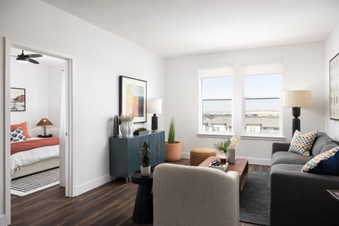 A living room with a grey couch, a grey armchair, a small table with a plant on it, and a bed with a red blanket in the background.