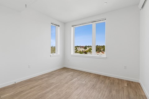 A room with white walls and wooden flooring, featuring two windows with a view of a residential area.