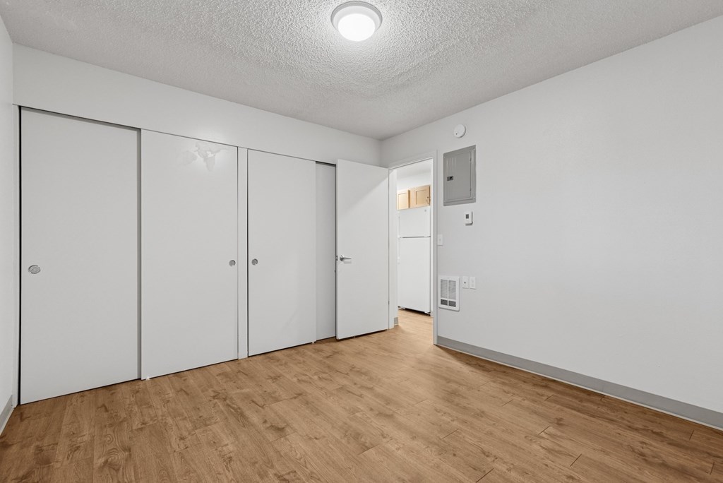 A room with white walls and wooden flooring, featuring a white door and a white closet with a mirror.
