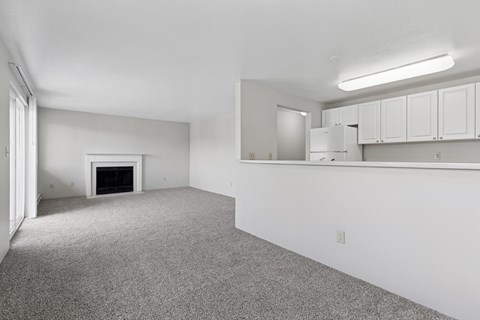 A sparsely furnished living room with a fireplace and a kitchen area in the background.