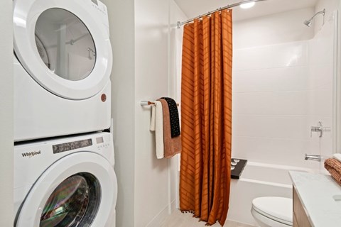 A white Whirlpool washing machine sits next to a bathroom with orange curtains.