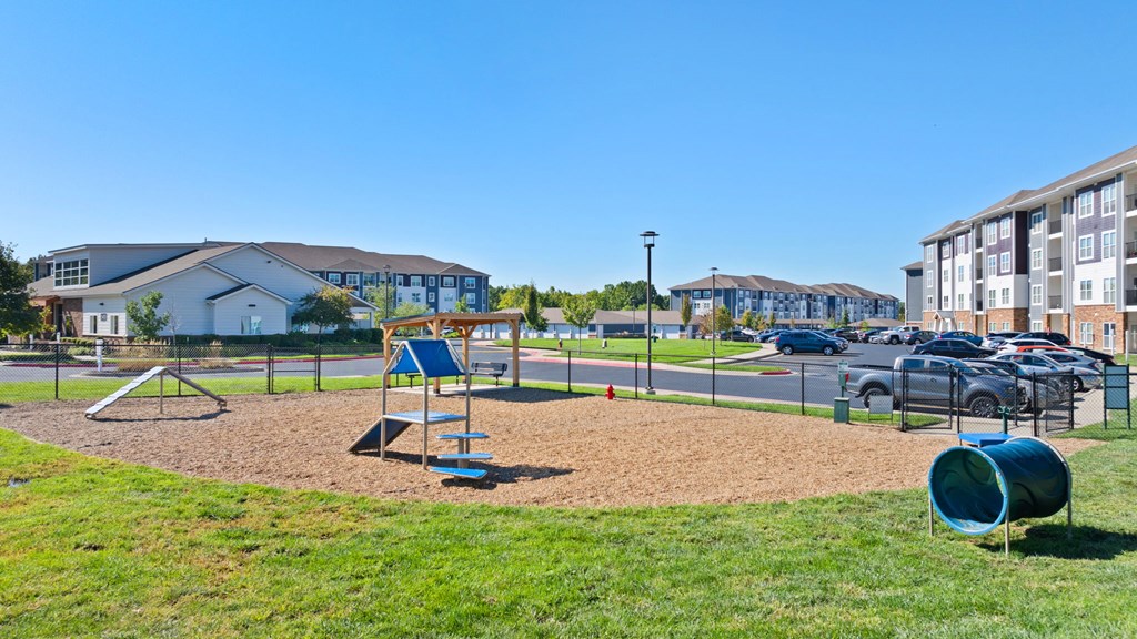 A playground with a slide, swings, and a seesaw in the foreground with a parking lot and buildings in the background.