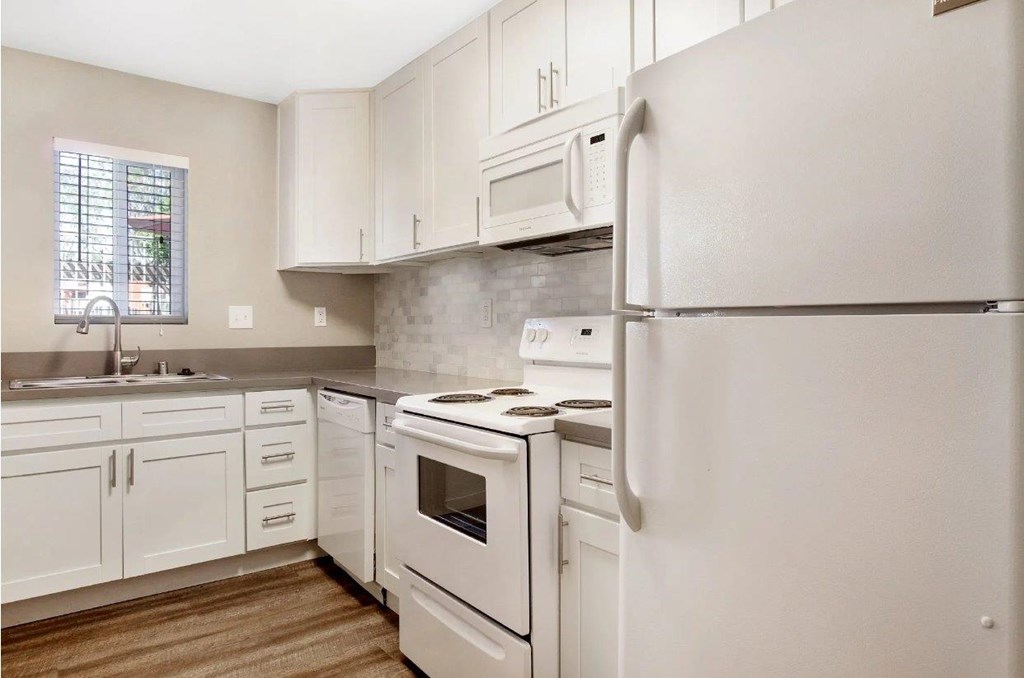 A white refrigerator stands in a kitchen with white cabinets and a white stove.