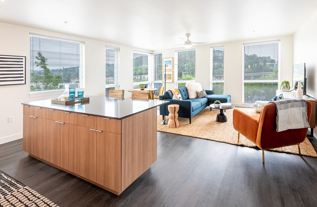 A large living room with floor to ceiling windows, dark wood plank flooring, breakfast bar, and an abundance of natural light.
