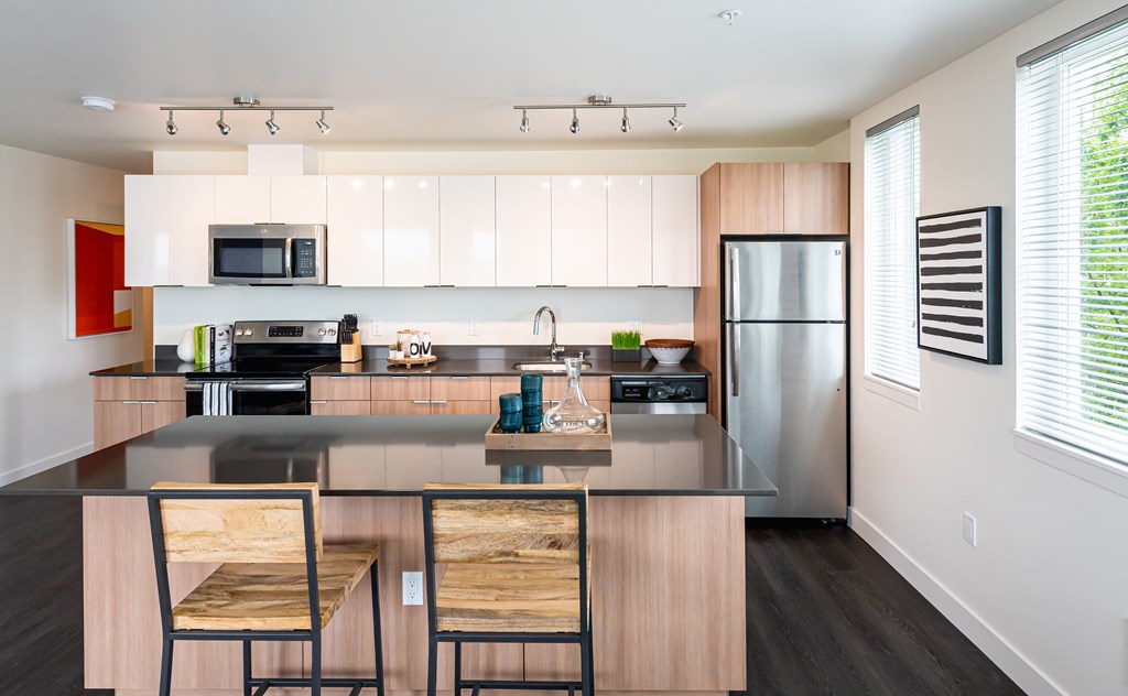 Stylish kitchen featuring white glossy upper cabinets and a light wood color lower cabinet. Black granite looking countertops, track lighting, stainless steel appliances, breakfast bar, vinyl wood flooring.