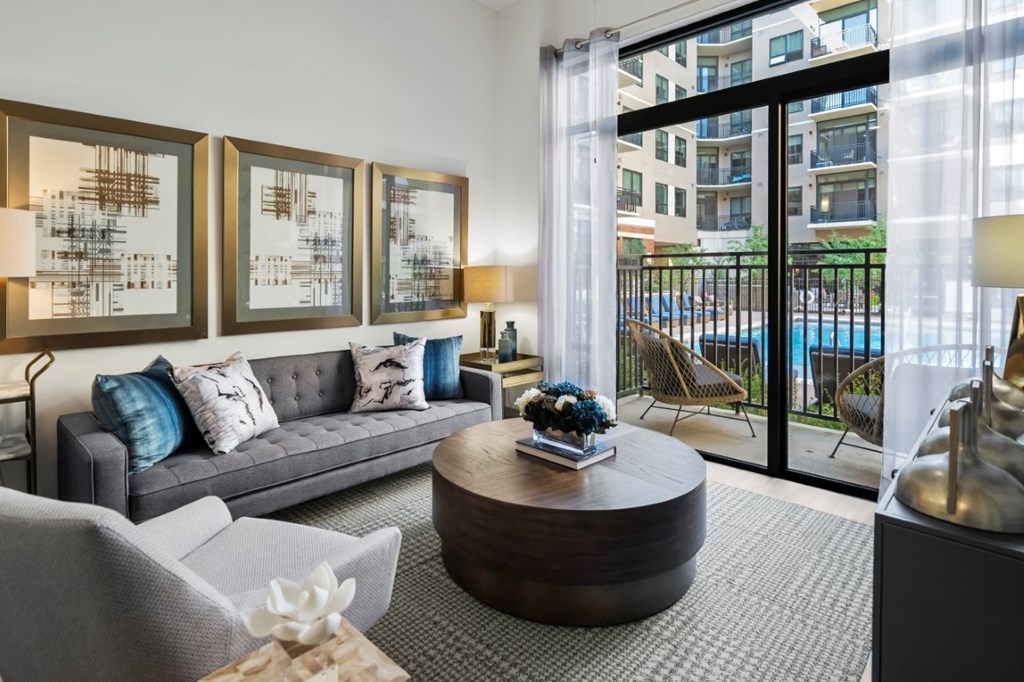 A living room with a grey couch, a coffee table, and a view of a pool and buildings outside.