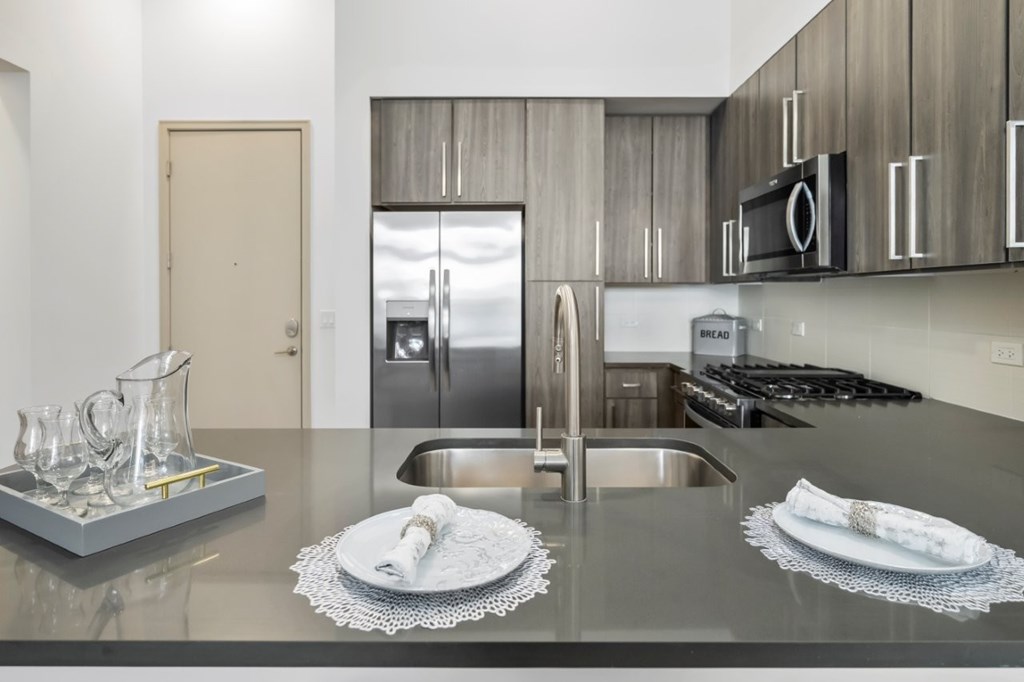 A modern kitchen with a stainless steel refrigerator, a sink, and a countertop with a tray of glasses and two plates.