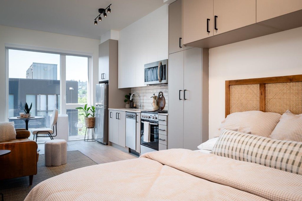 A modern bedroom with a large bed and a kitchen area in the background.