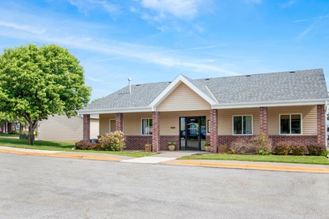 A small building with a grey roof and a black door is surrounded by a small grassy area and a paved road.