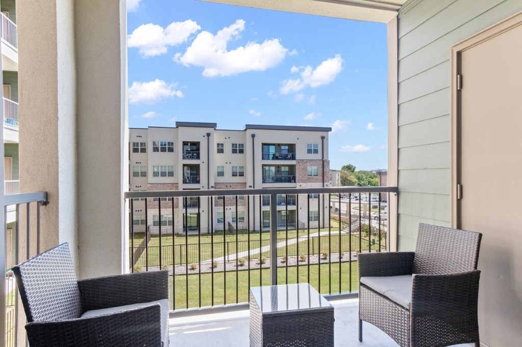 A balcony with two chairs and a table overlooks a parking lot and apartment buildings.