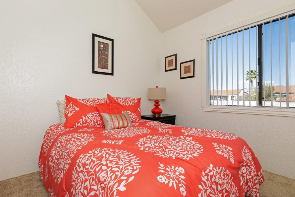 A bed with a red and white comforter is in the foreground of a room with a window.