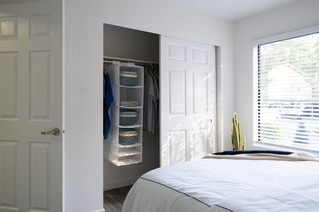 A white bedroom with a bed, a closet with clothes, and a window with blinds.