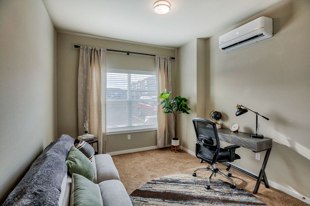 A room with a grey couch, a black chair, a desk with a lamp, and a window with curtains.