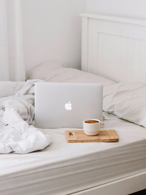 A laptop with an Apple logo is on a bed with a cup of coffee.