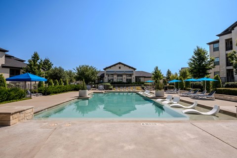A large swimming pool with a stone border and a stone patio with lounge chairs and umbrellas.