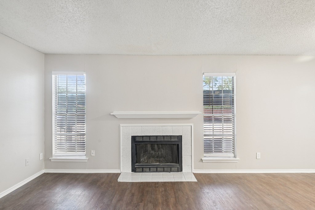 A white fireplace with a white mantle and a white base sits in a room with wood floors and two windows with blinds.