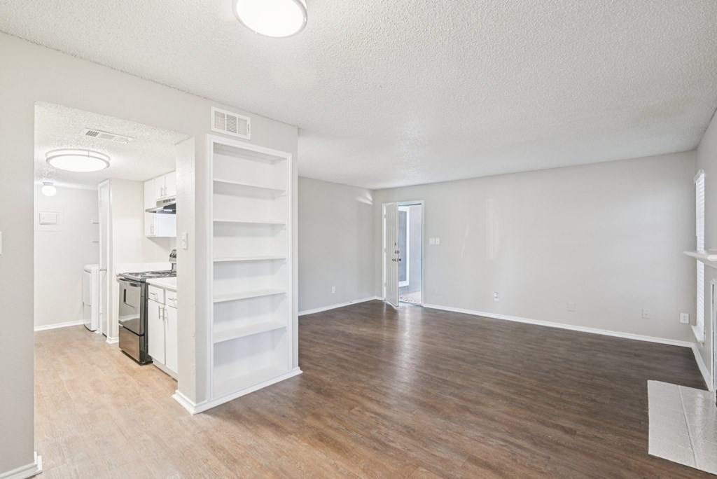 A white room with wood flooring and a kitchen area in the background.