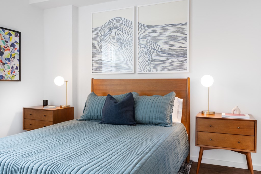 A bed with a blue comforter and two pillows sits in a room with a wooden nightstand and a painting on the wall.