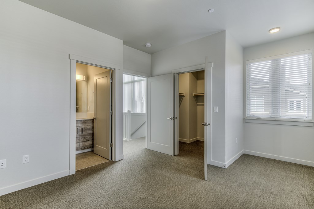 A spacious room with carpeted flooring and white walls, featuring a staircase and a window with blinds.