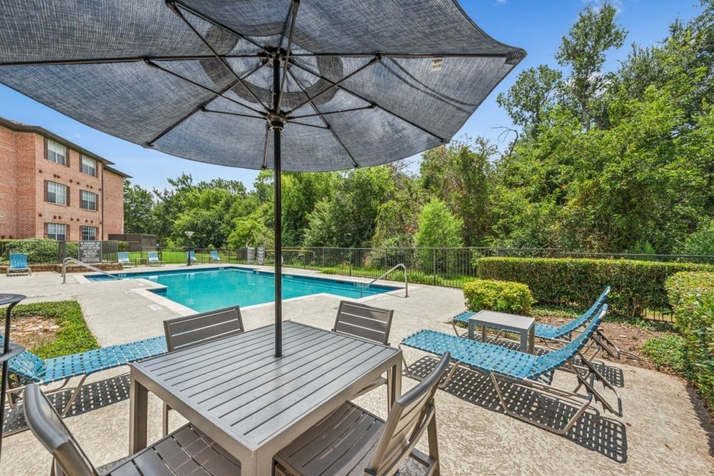 A table with chairs and an umbrella is in the foreground of a pool area.