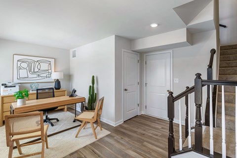 A room with a wooden desk and chairs, a cactus, and a staircase.