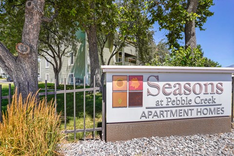 A sign for Seasons at Pebble Creek Apartment Homes is in front of a fence and trees.