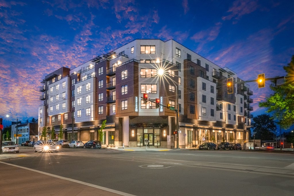 A modern apartment building is lit up at dusk with cars parked along the street.