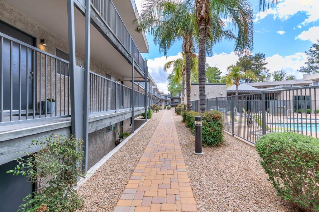 A long brick pathway leads through a landscaped area with a pool and palm trees.