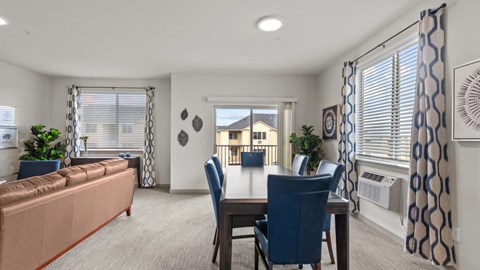 A living room with a brown sofa, a dining table with blue chairs, and a view of a building through the window.