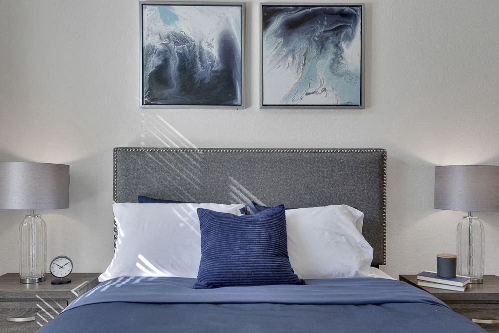 A bed with a blue comforter and two pillows with a grey headboard and two lamps on the side.