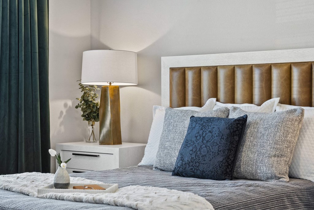 A bed with a white headboard and grey bedding with a lamp and vase on a nightstand.