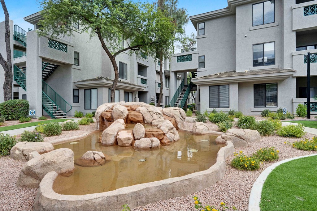 A small pond with rocks in the middle of a grassy area in front of apartment buildings.