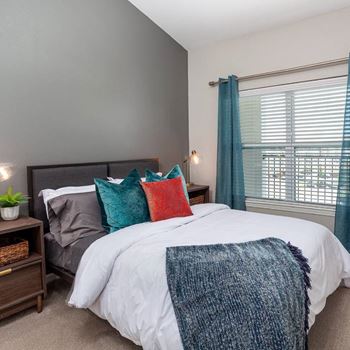 A bedroom with a white bed and a grey headboard, a blue blanket, and a red pillow.