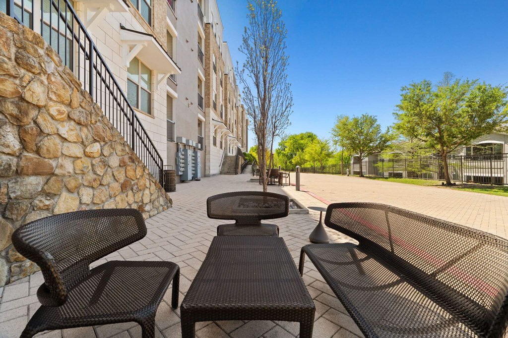 A stone wall separates a patio with three black chairs from a building.