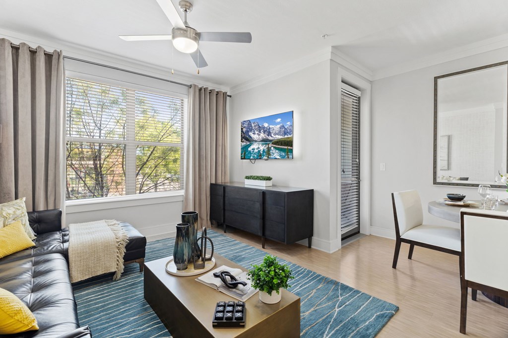 A living room with a black leather couch, a coffee table with a plant and a vase on it, a white chair, a dining table with a mirror, and a painting on the wall.