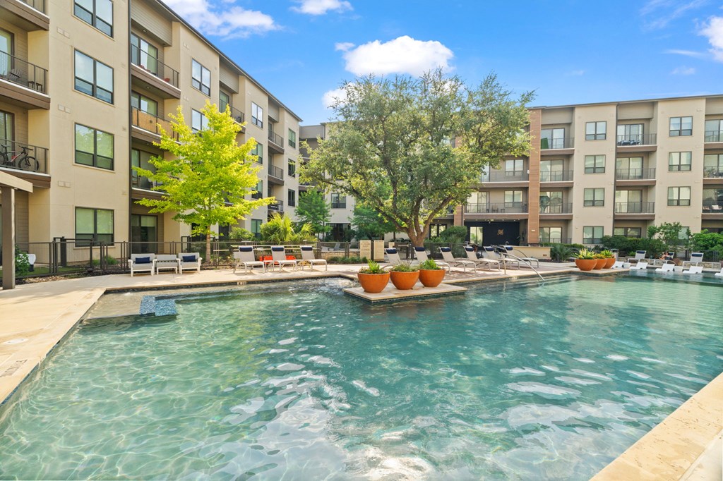 A swimming pool in front of apartment buildings with trees and lounge chairs.