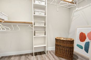 A white shelving unit with clothes hangers and a wicker basket in front of a painting.