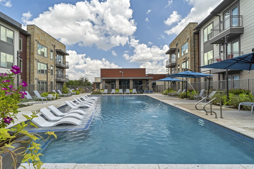 A swimming pool surrounded by lounge chairs and umbrellas in front of apartment buildings.